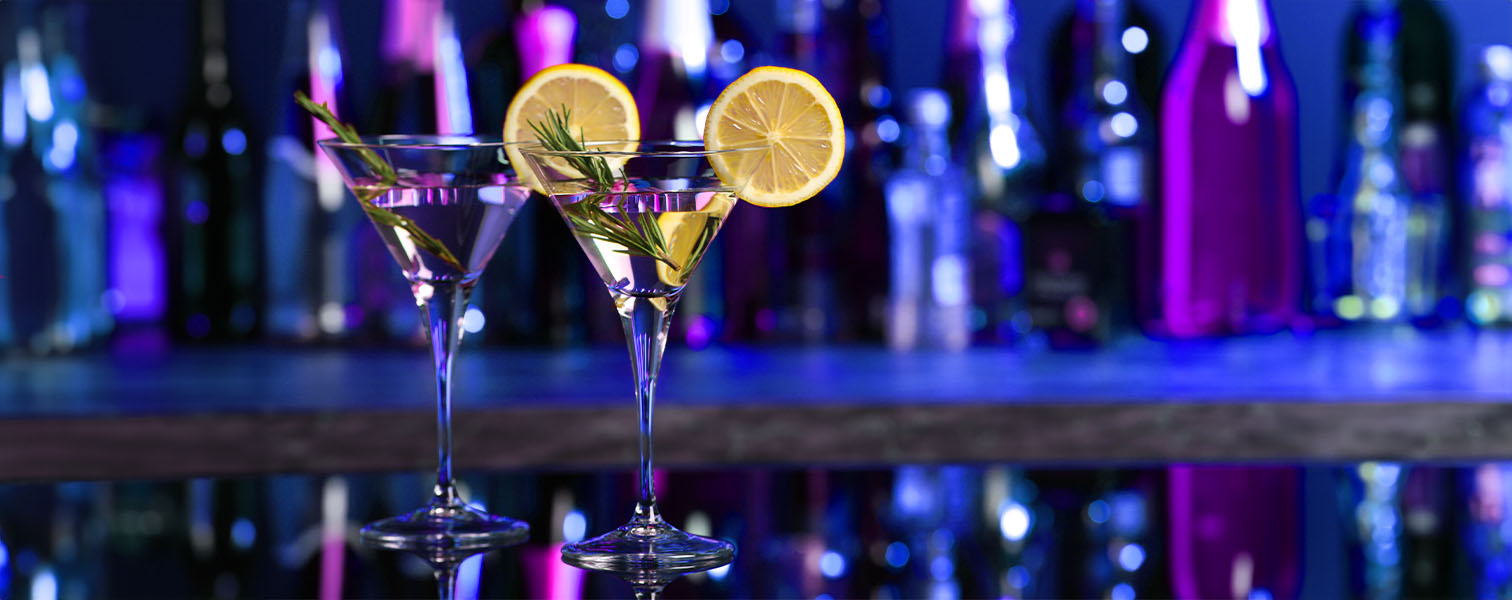 Two cocktails with rosemary and a lemon slice on a bar top with a purple blue ombre light in the background.