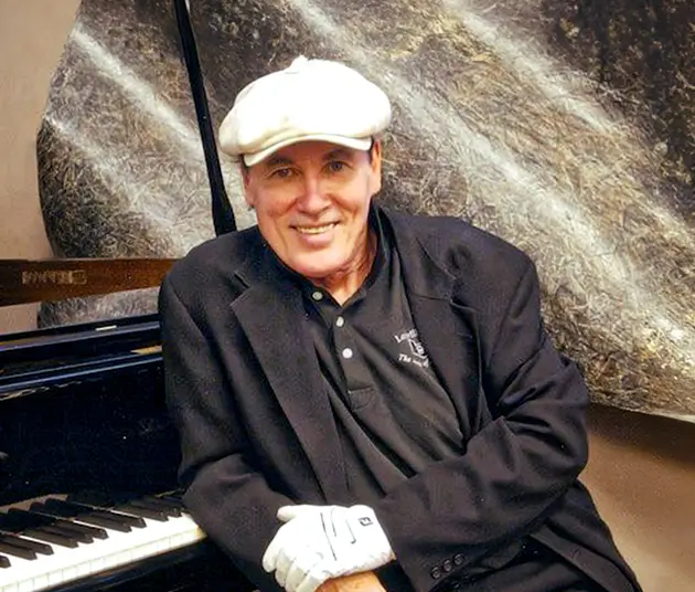 A picture of Billy Mac in a black suit jacket, black shirt, one white glove and a white hat next to a piano.