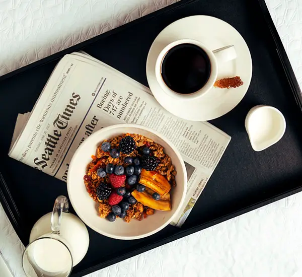 An image of a pitcher of milk, a bowl of granola and fruit, a Seattle Times newspaper, a cup of coffee with a rock sugar stick on the plate and a small cup of cream.