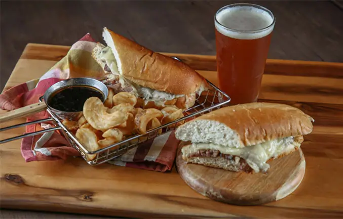 A burger with white cheese on a roll served with au jus, fries and a cold beer.