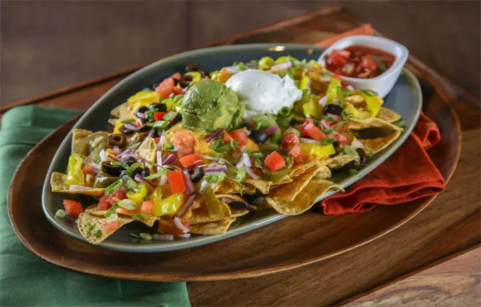 A plate of nachos topped with guacamole and sour cream served with a bowl of salsa.